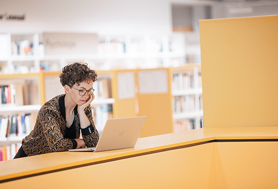 Verena Smit steht in einer Bibliothek vor ihrem geöffneten Laptop und arbeitet an einem Projekt der Wissenschaftskommunikation.